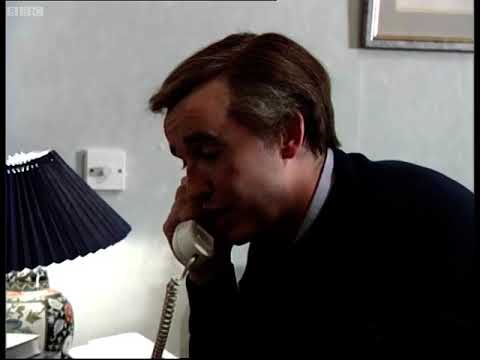 Alan Partridge taking a phone call while sitting on bed in a hotel bedroom. On the chest of draws in front of him is a portrait of David Hasselhoff