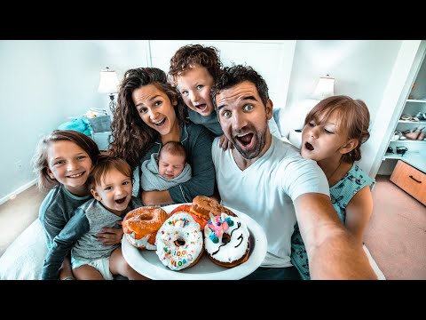 Family selfie with parents and kids gathered around a plate of colorful donuts, smiling in a bright home kitchen/room.