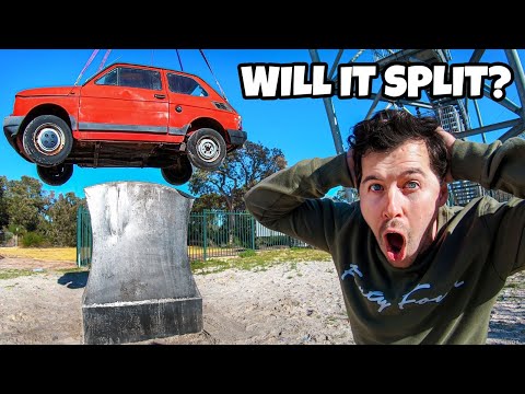 Man in a green sweatshirt with a shocked expression as a red car is hoisted by a crane above a concrete pedestal against a blue sky.