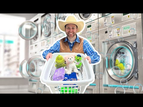 Smiling man in a cowboy hat and vest pushes a laundry basket filled with stuffed animals and laundry supplies in a laundromat.