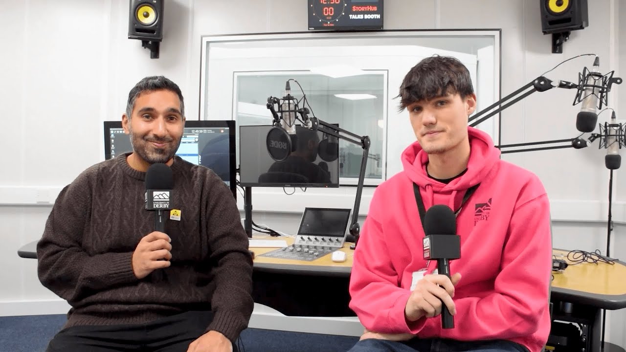 Baasit Siddiqui sits in a recording studio next to a student in a pink hoodie, they are both holding microphones.