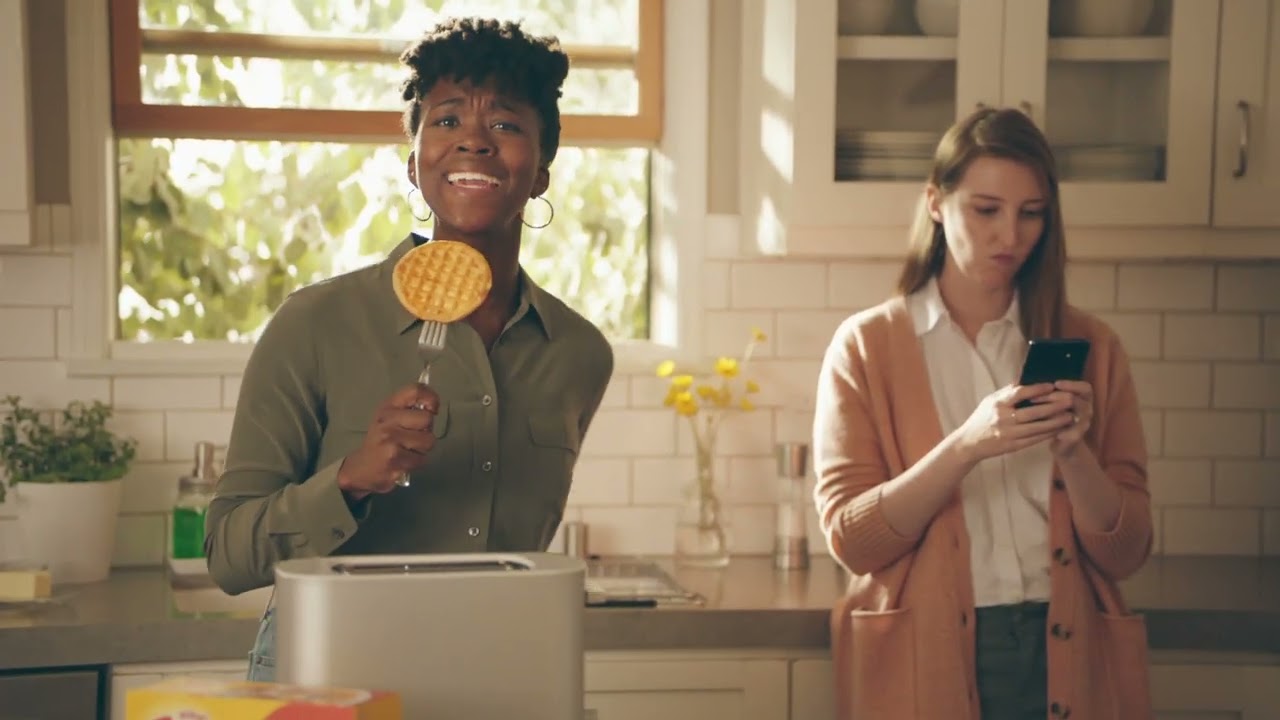 A woman holding an Eggo waflle like a microphone while another woman is looking at her phone