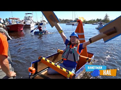 Woman in blue helmet and orange life vest cheers in a small DIY boat, cardboard wings raised above the water; harbor with boats and onlookers in the background.