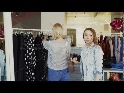 Young woman stealing wallet from female customer choosing clothes in shop. Thief looking around