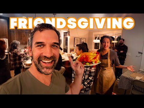 Selfie-style shot of friends at a kitchen party for Friendsgiving, a man in front smiles while presenting a plate of roasted turkey and sides to the group behind him.