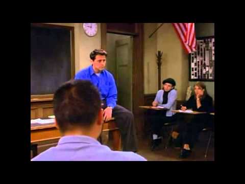 Joey from Friends TV show in a classroom sat casually on a desk at the front of a blackboard. Students are watching.