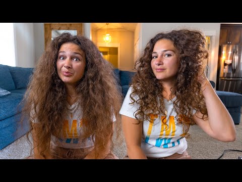 Two young women with long curly hair sit on the living room floor, smiling at the camera in a cozy home setting.
