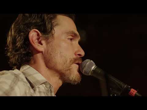 Trailer close-up: a singer at a lone mic, eyes closed, audience in silhouette