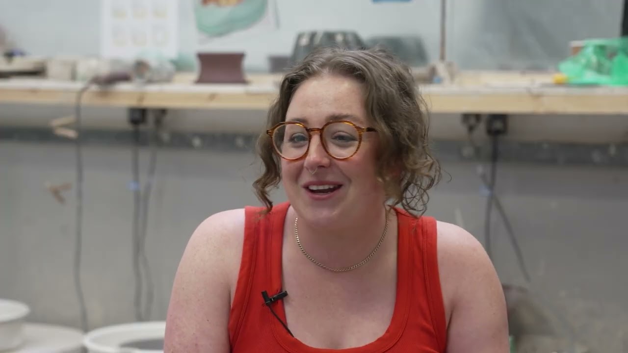 Alumni Abbie Sunter sits with her hair tied up loosely and wears an orange strappy top and glasses in a studio environment. 