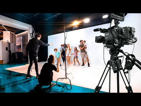 Behind-the-scenes of a studio photoshoot: kids posing in front of a white backdrop as crew adjust lighting and a large camera records nearby.