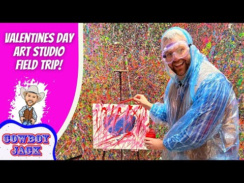 Man in a blue poncho and safety goggles paints a colorful splatter artwork on a small canvas, with a vibrant Valentine’s Day Art Studio backdrop behind him.