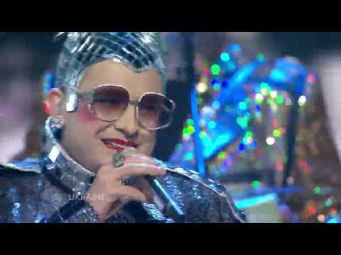 [[#Verka Serduchka [Βέρκα Σερντούτσκα]#]], "Dancing Lasha Tumbai" [Χορεύοντας λάσα τουμπάι], τελικός της Eurovision, Ελσίνκι 2007 (δεύτερη θέση). Το καμπ ως ολοκληρωμένο σύστημα υπερβολής: ο Ουκρανός κωμικός [[#Andriy Danylko [Αντρίυ Ντανίλκο]#]] εμφανίζεται με τη ντραγκ περσόνα του, μια μεσήλικη συνοδό κλινάμαξας από την Πολτάβα, ντυμένος ντισκομπάλα, με υπερμέγεθες ακορντεόν και χορευτές με κούρεμα μαλλέ. Η στυλιζαρισμένη οικειοποίηση του «κακού μετασοβιετικού γούστου», η υβριδική γλώσσα σουρζίκ και η εναρκτήρια ατάκα «me English nicht verstehen. Let's speak dance» μετατρέπουν το στίγμα της επαρχιακής αμορφωσιάς σε επιτελεστικό κεφάλαιο, ενώ η φανταχτερή θηλυκότητα παραμένει «ασεξουαλική», σύμφωνα με την κατά Sontag έννοια του καμπ σώματος. Τα ανύπαρκτα «lasha tumbai», που η επιτελέστρια απέδιδε ως μογγολική λέξη για τη σαντιγ;y, ακούστηκαν ως «Russia goodbye» και προσέδωσαν στο νούμερο πολιτικό διφορούμενο. Η αμφιθυμία της υποδοχής επιβεβαιώνει τη λειτουργία του καμπ: εικόνες της κάηκαν στο Μαϊντάν, βουλευτές την αποκάλεσαν γκροτέσκα και χυδαία, και ταυτόχρονα κέρδισε το βραβείο Barbara Dex για τη χειρότερη αμφίεση, διάκριση που στη λογική του είδους ισοδυναμεί με έπαινο.