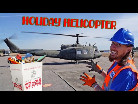 Volunteer in a blue hard hat and orange vest beside a U.S. Army helicopter on a tarmac, with a Toys for Tots box in front.