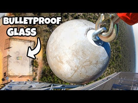 Drone view of a large round ballast sphere attached to a crane on a high-rise balcony, with 'BULLETPROOF GLASS' text overlay