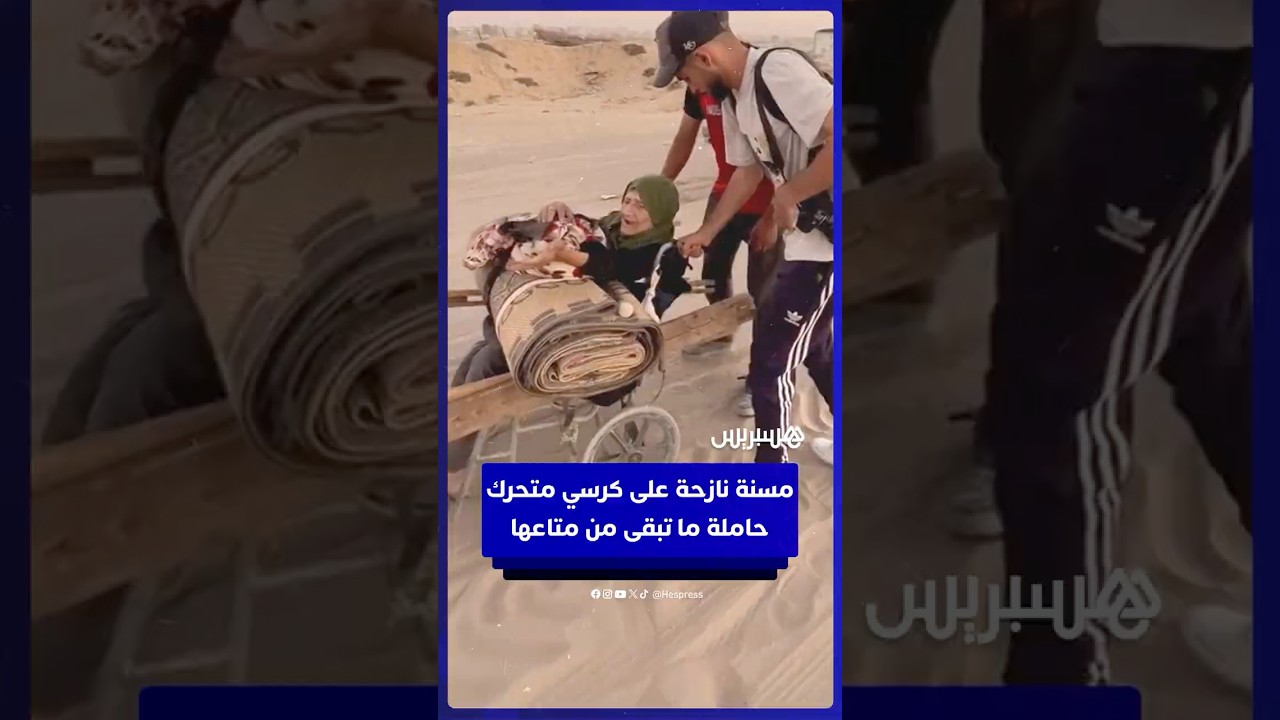 مسنة فلسطينية تتنقل على كرسي متحرك حاملة ما تبقى من متاعها بينما يمدّ لها الشباب يد العون على الطريق thumbnail