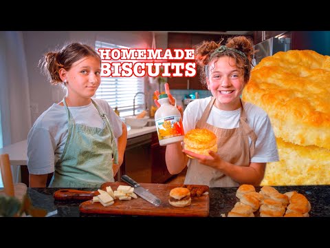 Two girls in aprons in a kitchen proudly display a homemade biscuit and a bottle of maple syrup by a cutting board with butter and a knife visible.