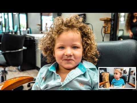 Child with curly hair sits in a barber chair, smiling at the camera in a salon setting