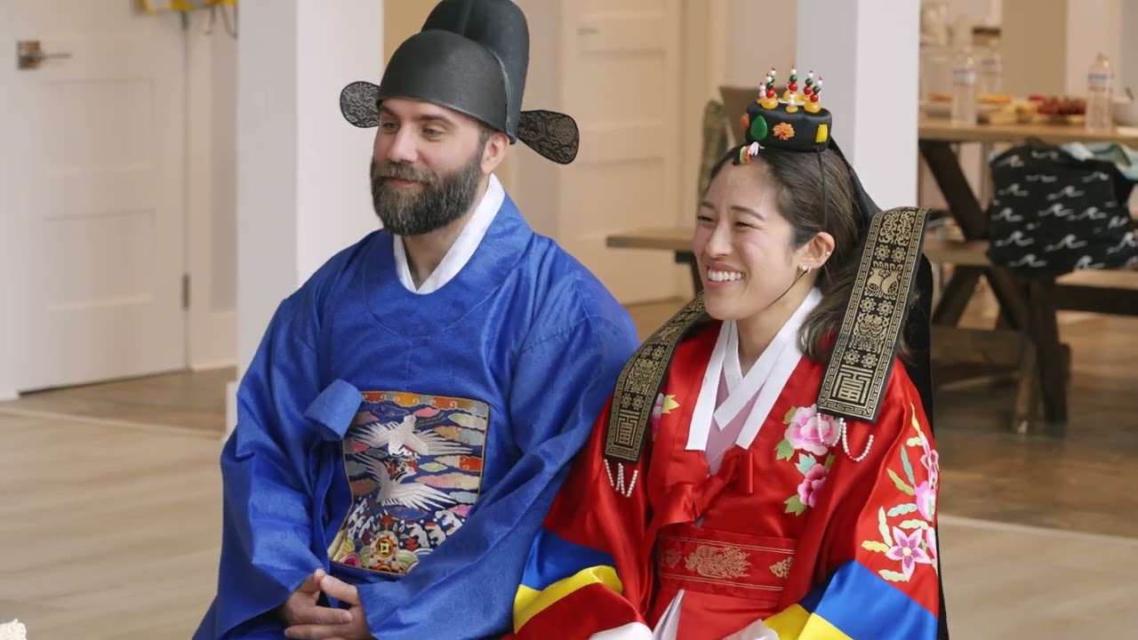 A Korean bride and groom in colorful hanbok bowing to family members during a traditional pyebaek tea ceremony with dates and chestnuts on a low table