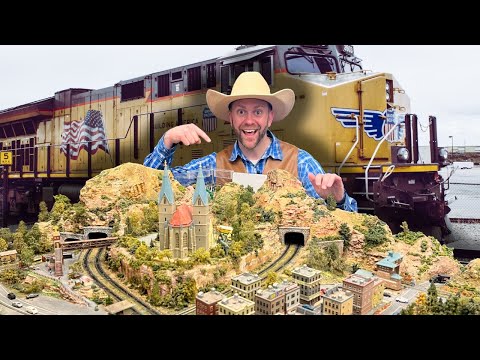 Man in a cowboy hat and blue plaid shirt points to a detailed model railroad landscape with tunnels and a church, set in front of a large yellow locomotive behind him.
