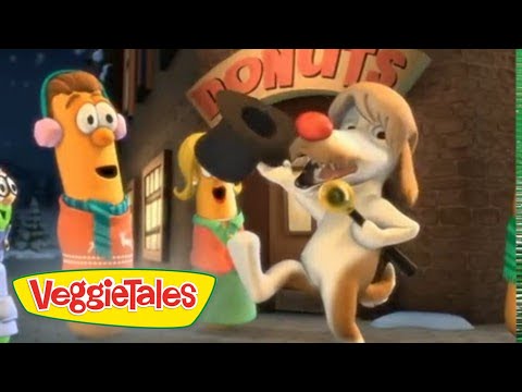 Animated VeggieTales scene: a white dog with a red nose rings a bell while a carrot character watches nearby near a building with VeggieTales signage in the background.