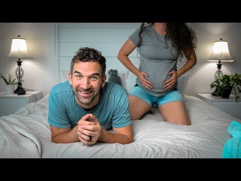 Smiling man lying on a bed with a pregnant woman kneeling behind him, hands on her baby bump in a cozy, lamp-lit bedroom.