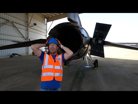 Maintenance worker in a high-visibility vest and hard hat poses with hands behind head beside a jet engine inside an aircraft hangar.