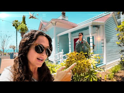 Woman wearing sunglasses takes a selfie in front of a bright turquoise house, while a man in a green flight suit stands on the porch in the background under a sunny sky.