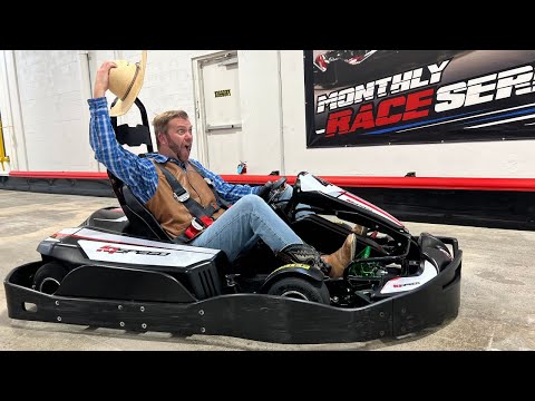 Man in a go-kart raising a straw hat with an excited expression indoors, wearing a blue plaid shirt and brown vest.