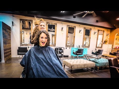 Woman seated in a salon cape while a stylist lifts her hair from behind, smiling at the camera in a modern hair salon setting.