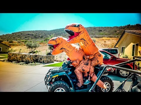 Two people in orange inflatable dinosaur costumes riding an ATV in a sunny residential driveway.