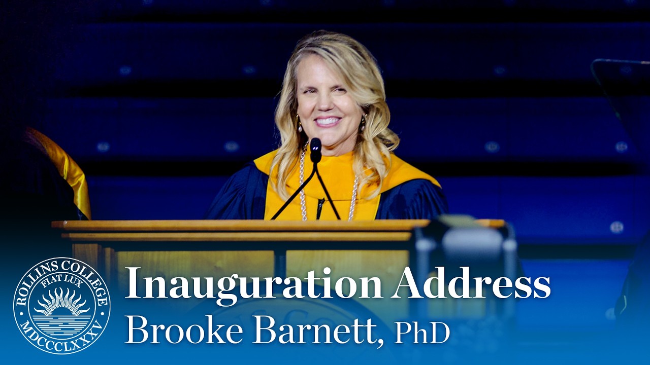 ��ɫ�� President Brooke Barnett stands on stage in academic regalia at the presidential inauguration with a college seal in the background.