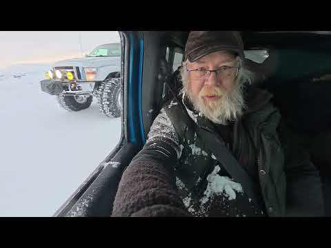 Photograph of a bearded man in winter gear sitting in a vehicle, with a truck visible through the window in a snowy landscape.