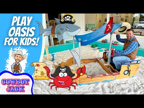 Man in a blue plaid shirt and brown vest sits in a carved wooden pirate ship sandbox with a blue canopy and cartoon decorations for kids activity.
