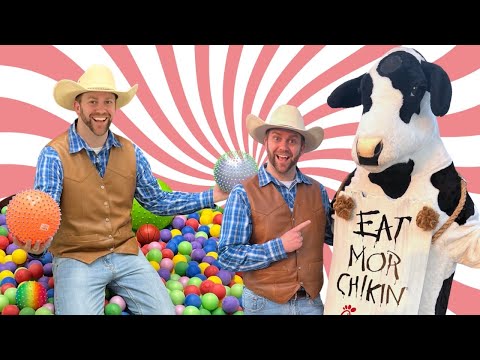 Two men in cowboy vests and hats pose in a colorful ball pit with a life-size cow mascot holding a sign that says 'EAT MOR CHIKIN' against a pink swirl background.