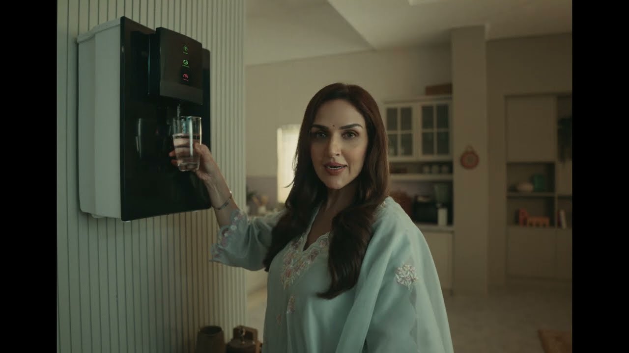 A woman in a pale blue kurta fills a glass at a wall-mounted water dispenser