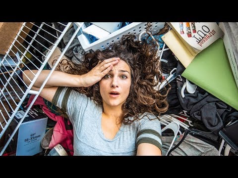 Woman lying on her back amid a chaotic pile of belongings, holding her forehead in surprise or worry, eyes wide.
