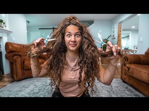 Woman sitting on the floor, holding scissors in each hand and teasing her long curly hair with a playful expression in a living room.