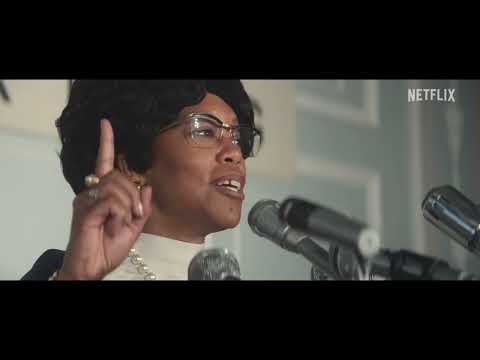 Trailer punch-in: Chisholm at a podium, brass buttons catching light as the strings tighten