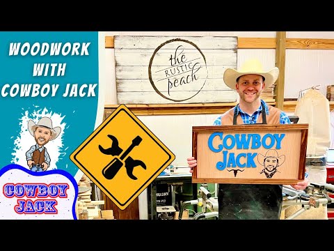 Smiling man in a cowboy hat holds a wooden 'Cowboy Jack' sign in a busy woodshop.