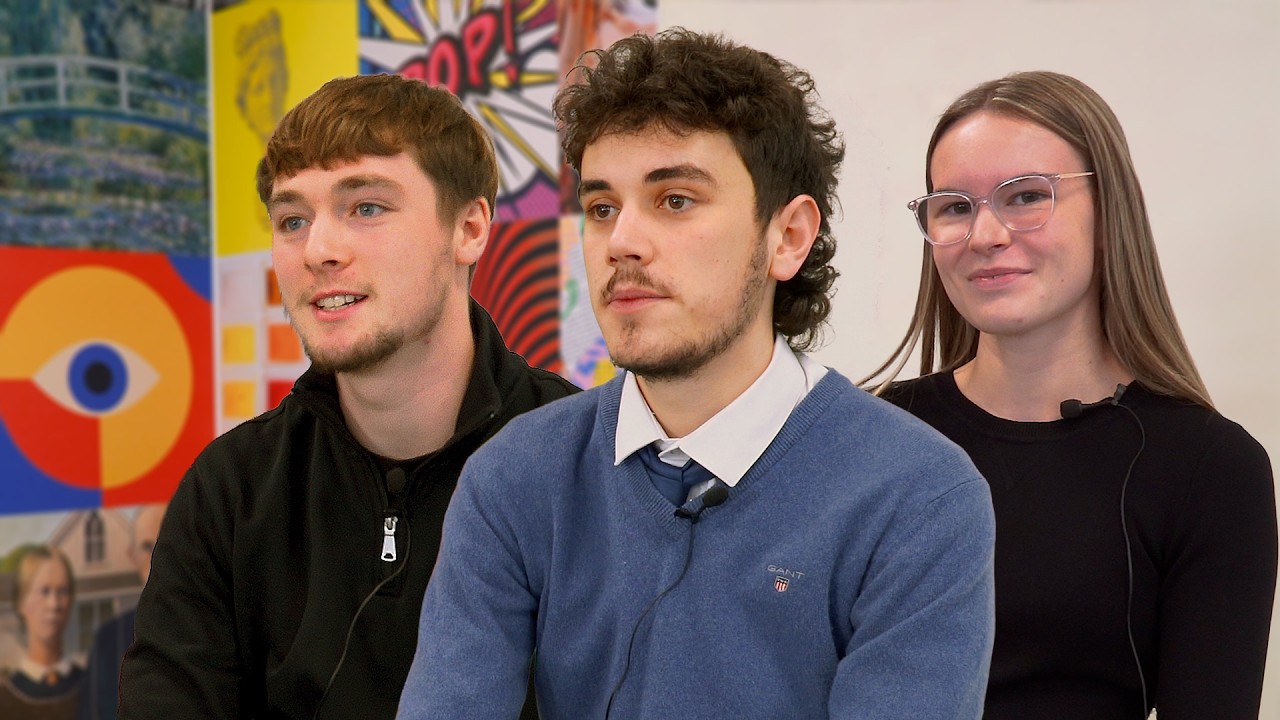 A group of three males in hoodies and casual attire stand with microphones with a busy classroom environment behind them.