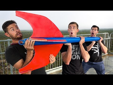 Three men on a balcony hold a oversized red and blue prop with surprised expressions on their faces.