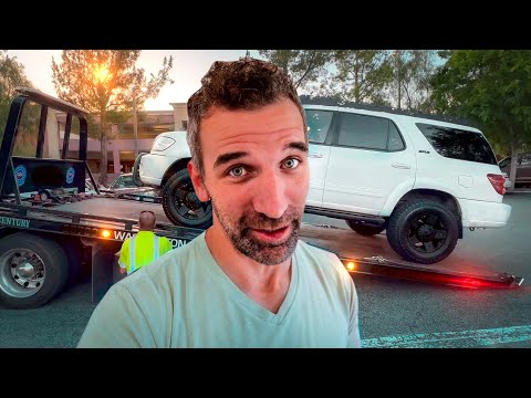 Man takes a selfie in front of a tow truck loading a white SUV onto a flatbed at sunset in a parking lot.