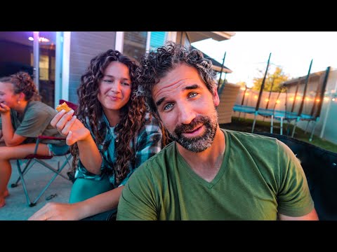 A man in a green shirt leans toward the camera while a smiling woman with curly hair holds a cracker beside him in a backyard setting with a trampoline in the background.