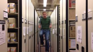 A student standing sitting on a step ladder between two rows of bookshelves.