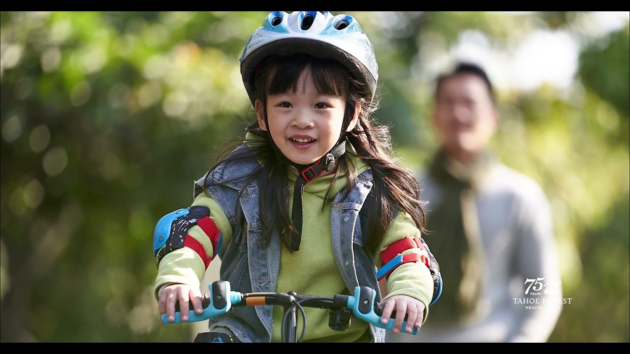 Young girl wearing a helmet riding a bicycle with her father in the blurred background