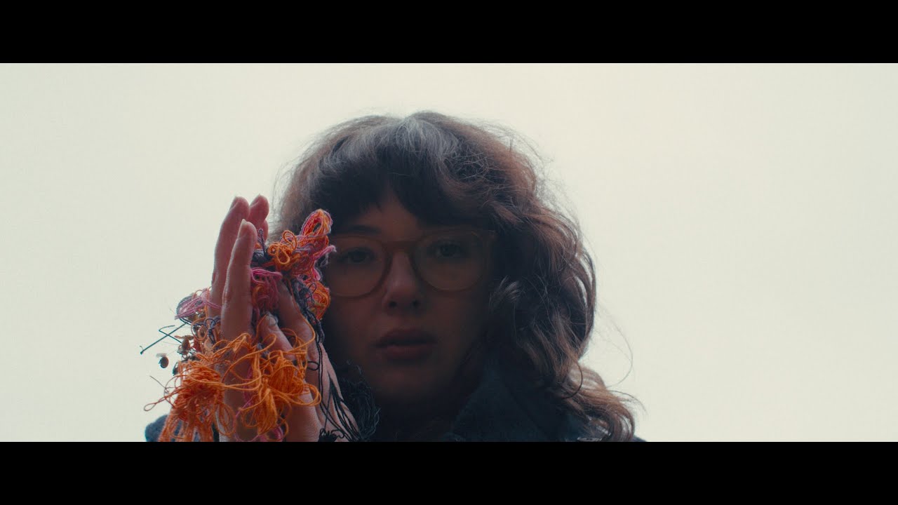 A moody, cinematic film still. Close-up of intertwined hands in low warm candlelight against near-black background, burgundy and amber tones.