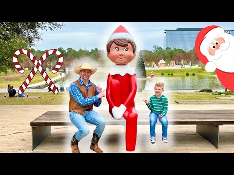 Man in a cowboy hat and vest sits on a bench beside a large red elf statue while a smiling boy sits on the other end, in a Christmas-decorated park.