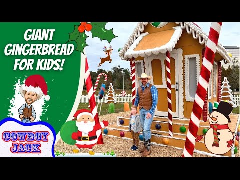 Father and child in cowboy outfits stand beside a giant gingerbread house at a holiday display with large candy canes and festive decorations.