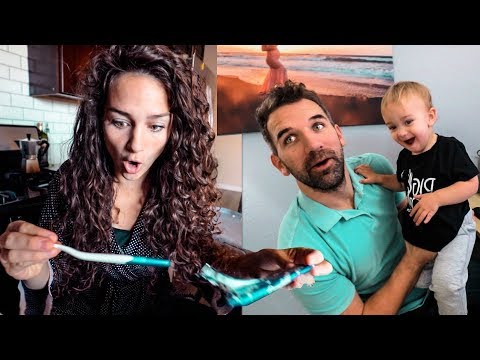 Woman with curly hair looks surprised while holding a turquoise item toward the camera; a man holds a smiling baby beside her in a kitchen/dining area.