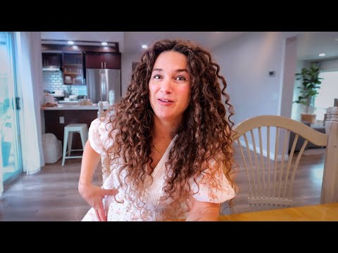 Woman with long curly hair sits at a wooden dining table in a bright kitchen, smiling at the camera. (Informative, concise)